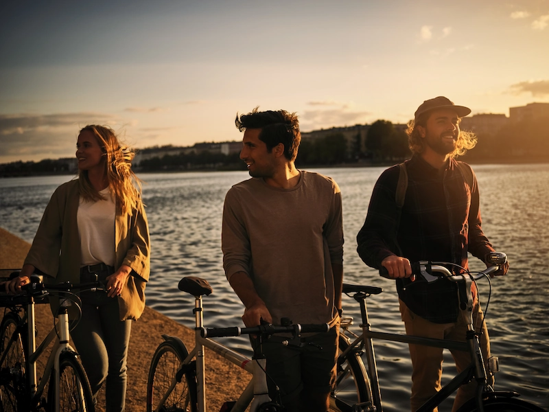 Drie jongvolwassenen staan met hun fietsen bij het water tijdens zonsondergang.
